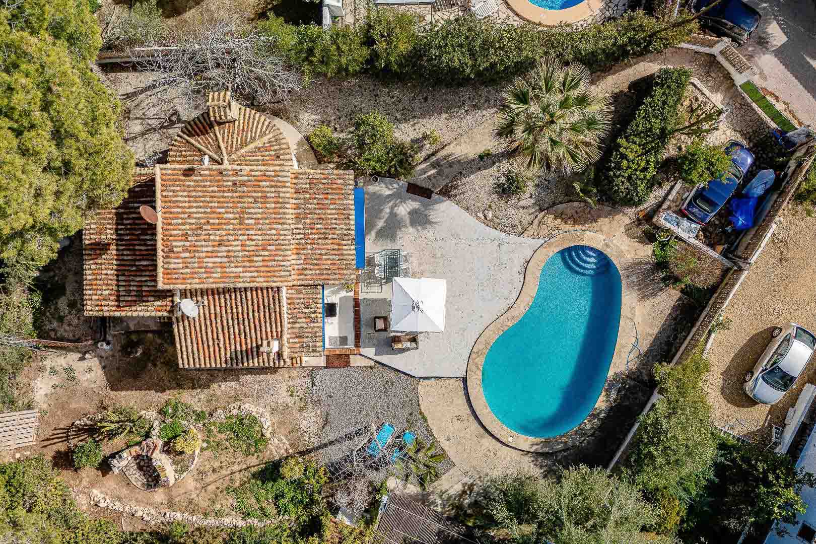Vista aérea cenital de la villa con piscina de forma riñonada, terraza pavimentada y tejado de teja tradicional rodeado de vegetación mediterránea.