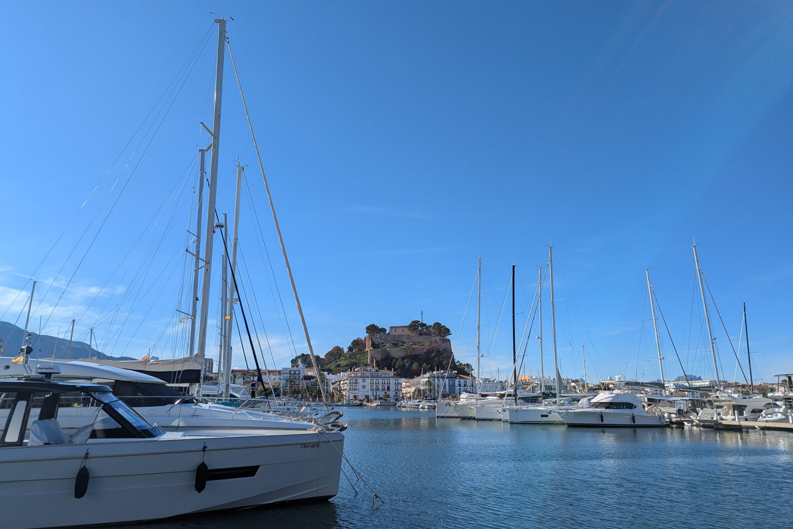 Vista del puerto deportivo con yates y el castillo de Dénia al fondo bajo un cielo azul despejado.
