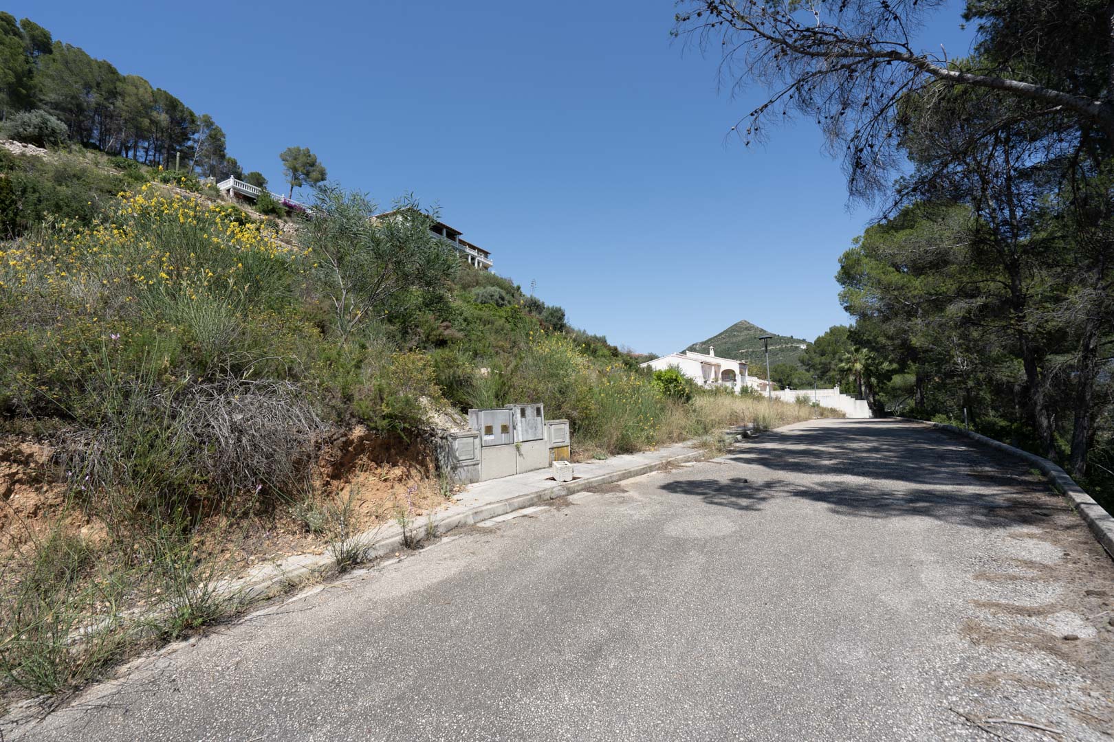 Acceso por carretera asfaltada al Residencial VALL DE POP en Alcalalí, mostrando la infraestructura de suministros y el entorno natural de la ladera.