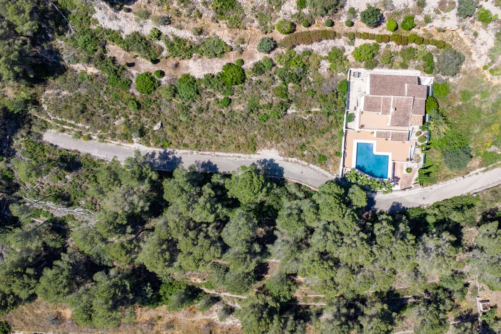Vista aérea cenital de la villa integrada en la ladera boscosa de Alcalalí, destacando la piscina privada en forma de L y el acceso por camino sinuoso.