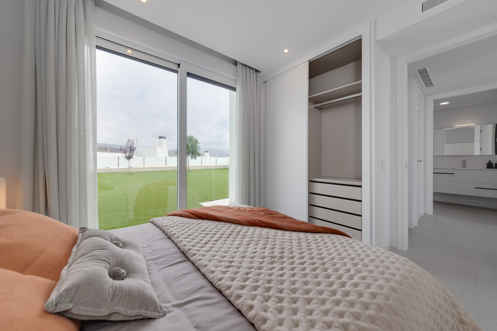 Dormitorio con armario empotrado de puertas correderas, grandes ventanales con acceso directo al jardín y vista hacia el baño en suite con mobiliario suspendido.