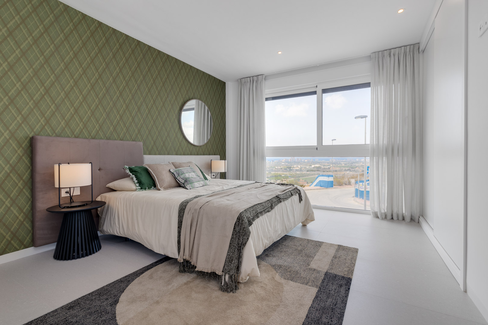 Dormitorio con pared decorativa en verde, ventanal de suelo a techo con vistas despejadas y armario empotrado blanco. Incluye iluminación empotrada y suelos porcelánicos.
