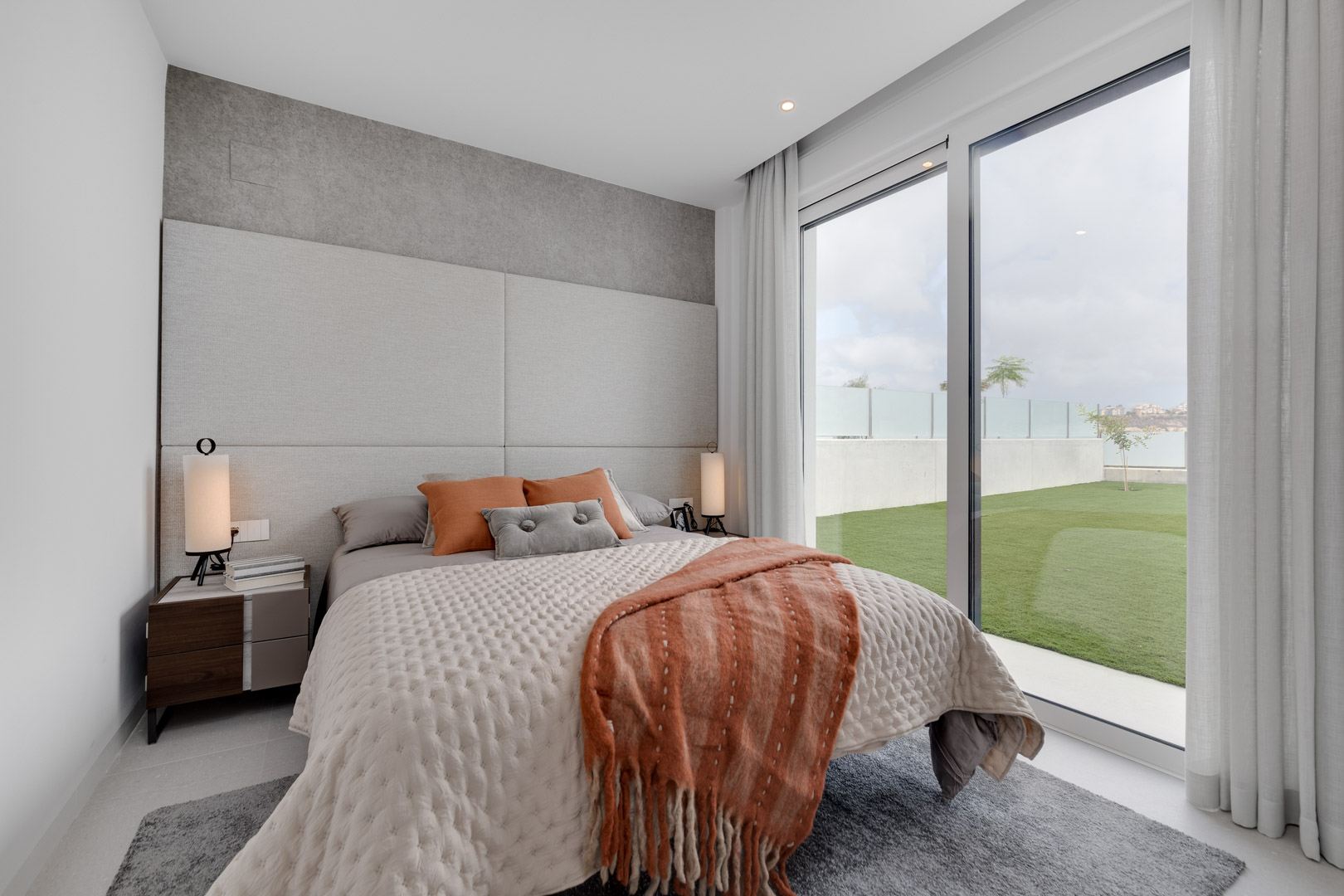 Dormitorio contemporáneo con cabecero tapizado, textiles en tonos tierra y grandes ventanales con acceso directo a la zona de jardín privado.