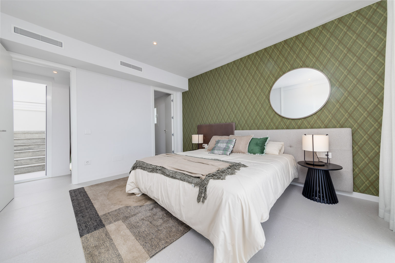 Dormitorio contemporáneo con pared de acento en papel pintado verde oliva, espejo circular y sistema de climatización integrado mediante rejillas lineales.