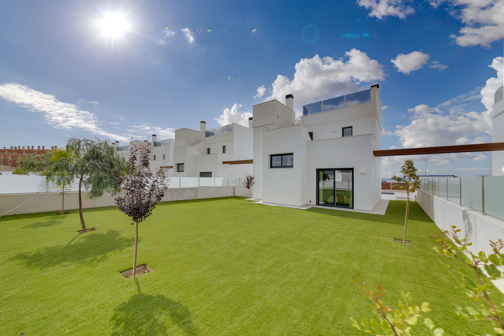 Exterior de la villa con fachada blanca minimalista, amplio jardín de césped y árboles jóvenes bajo un cielo despejado.