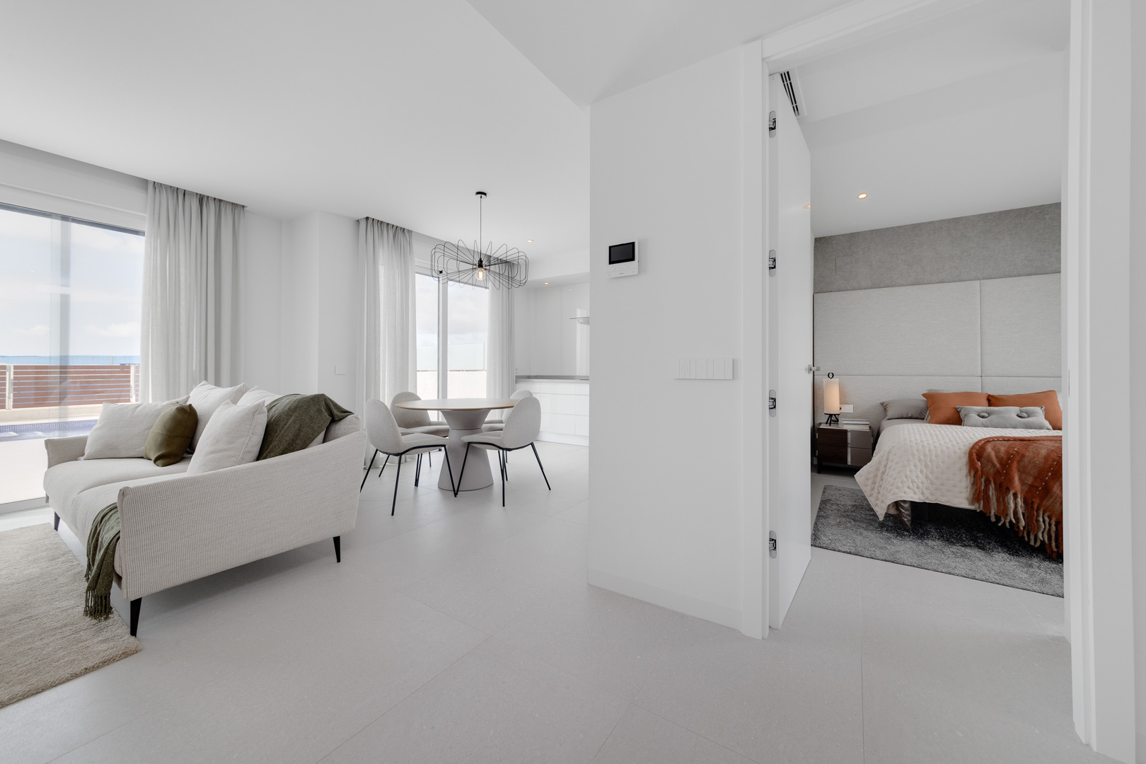 Salón-comedor de concepto abierto con suelos porcelánicos, mobiliario contemporáneo y acceso visual a un dormitorio con textiles en tonos tierra y cabecero tapizado.