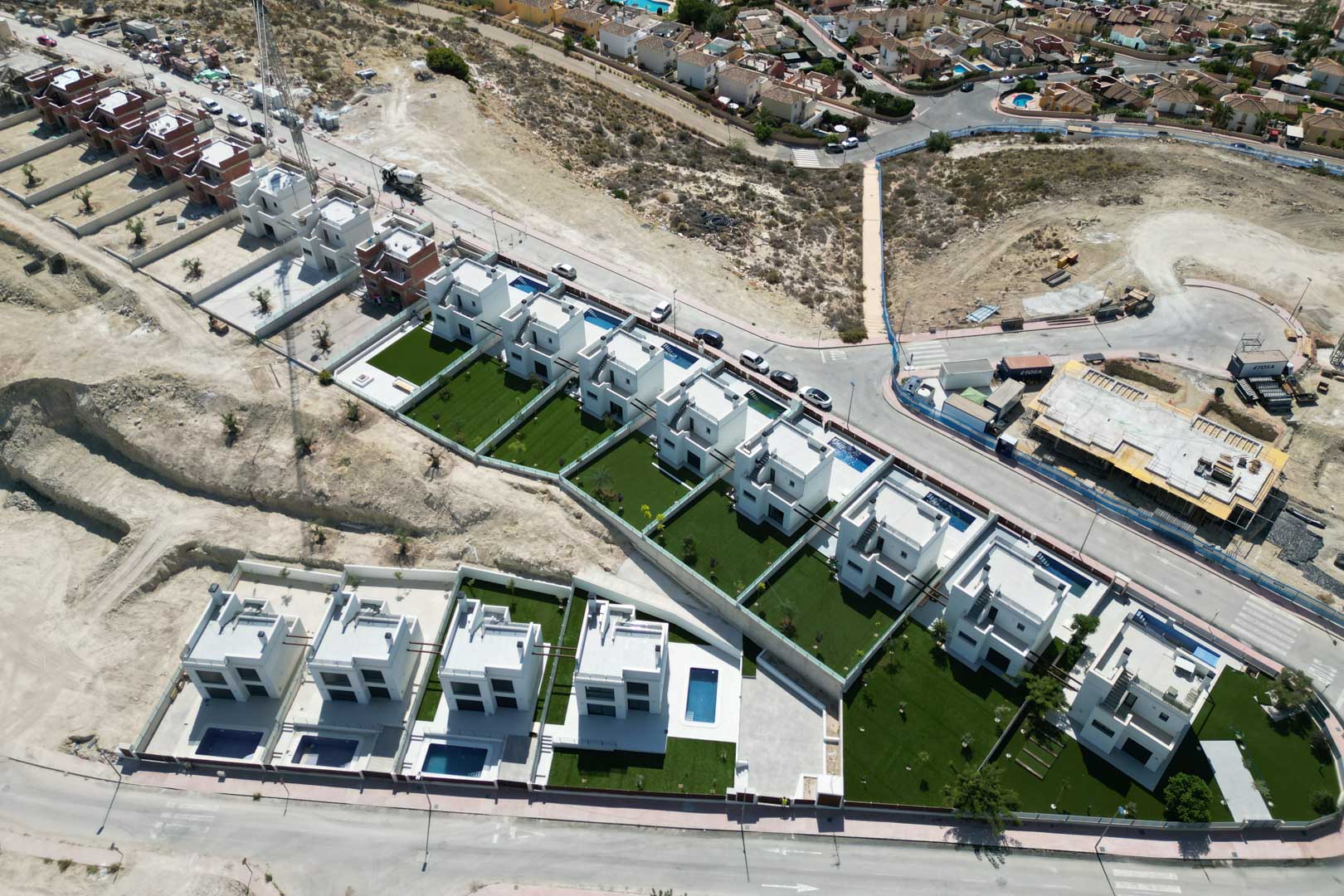 Vista aérea de una promoción de villas modernas blancas con piscinas privadas y jardines, situadas en una zona elevada en fase de desarrollo.