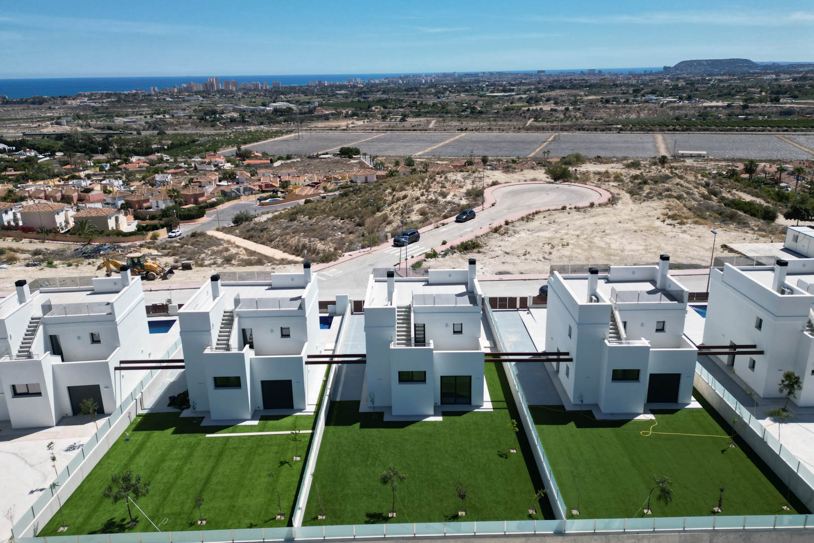 Vista aérea de villas contemporáneas con fachadas blancas, jardines privados de césped y terrazas superiores, situadas frente a una panorámica del mar y la costa.