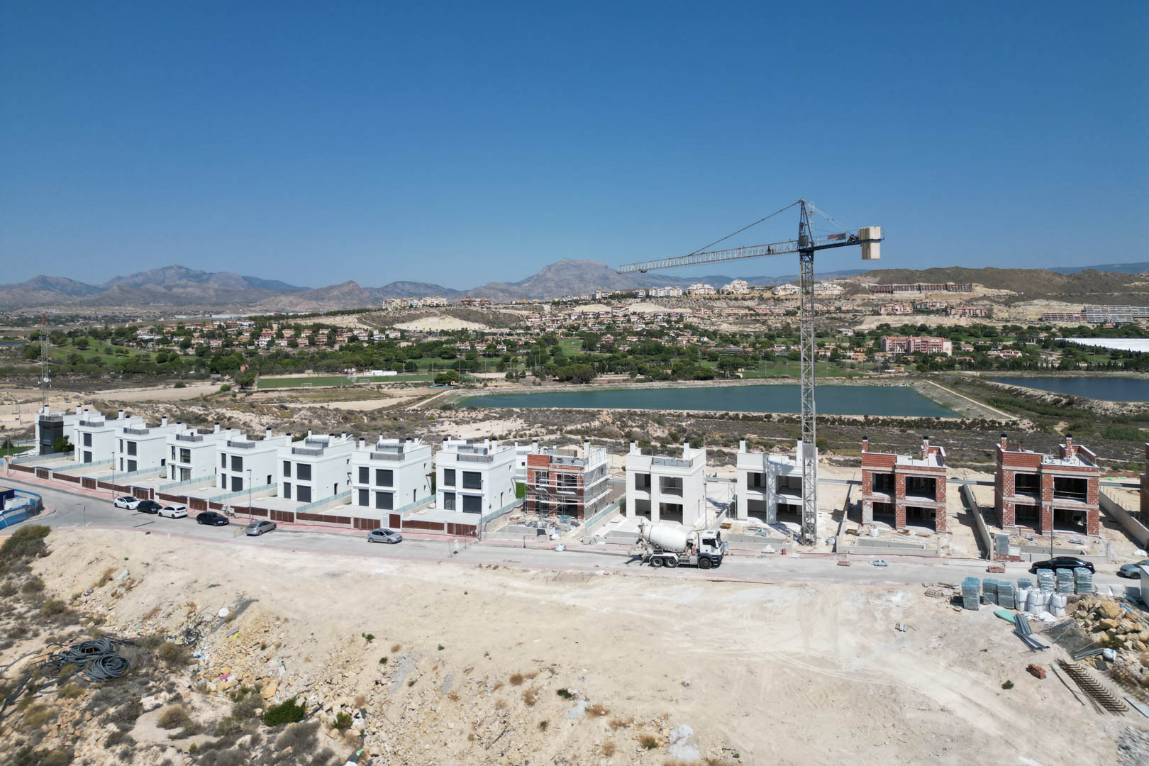 Vista aérea del progreso de construcción de las villas contemporáneas, mostrando las fachadas blancas terminadas y estructuras en obra frente al campo de golf y las montañas.