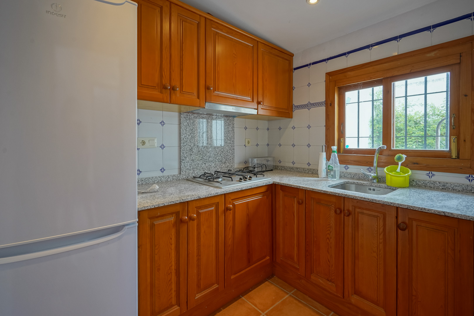 Cocina funcional con armarios de madera maciza, encimera de granito gris y placa de gas. Incluye fregadero bajo ventana de madera y suelos cerámicos tradicionales.