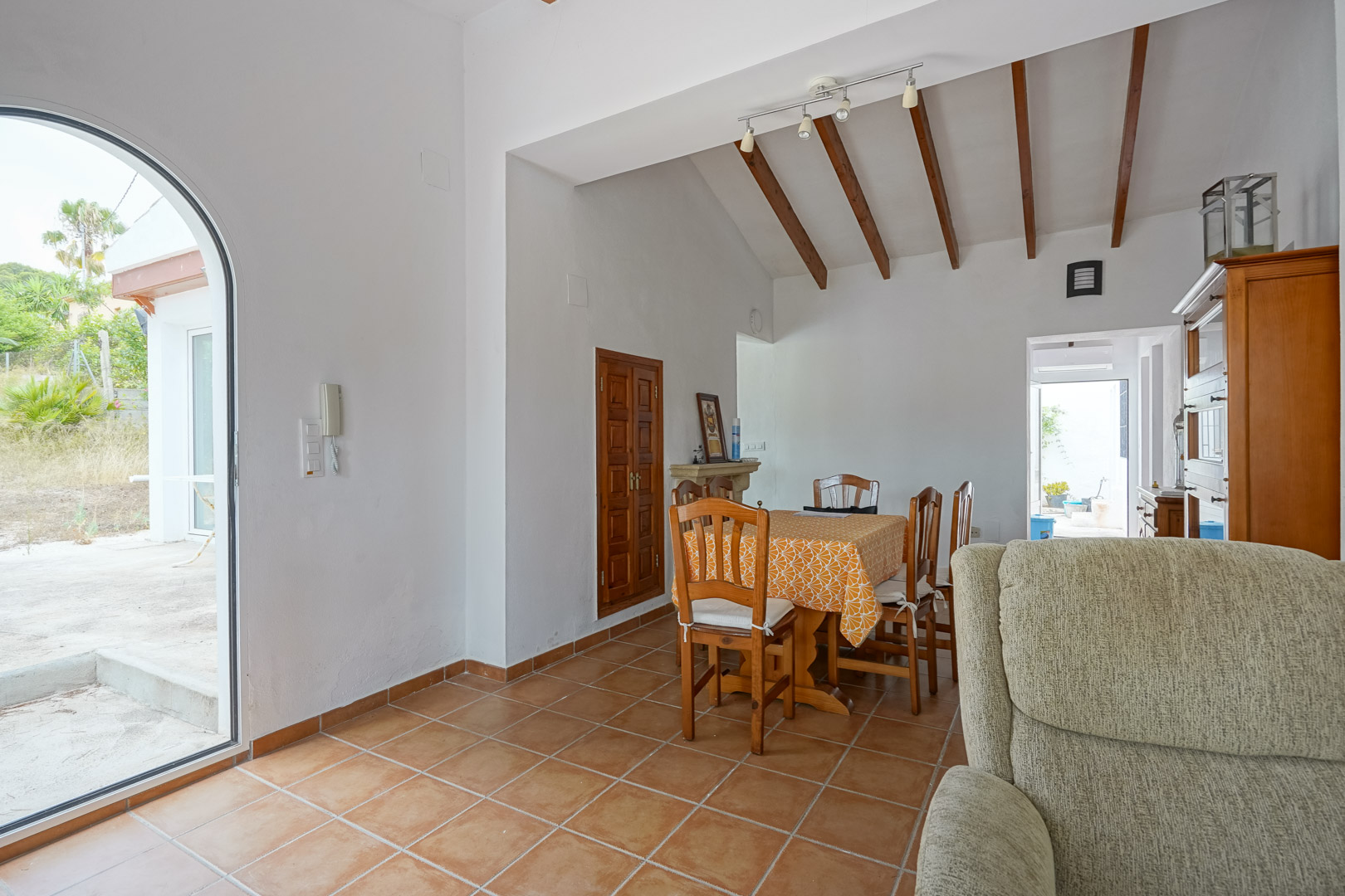 Comedor con techos altos de vigas de madera vista, suelos de gres rústico y acceso directo al exterior mediante una puerta de arco acristalada.
