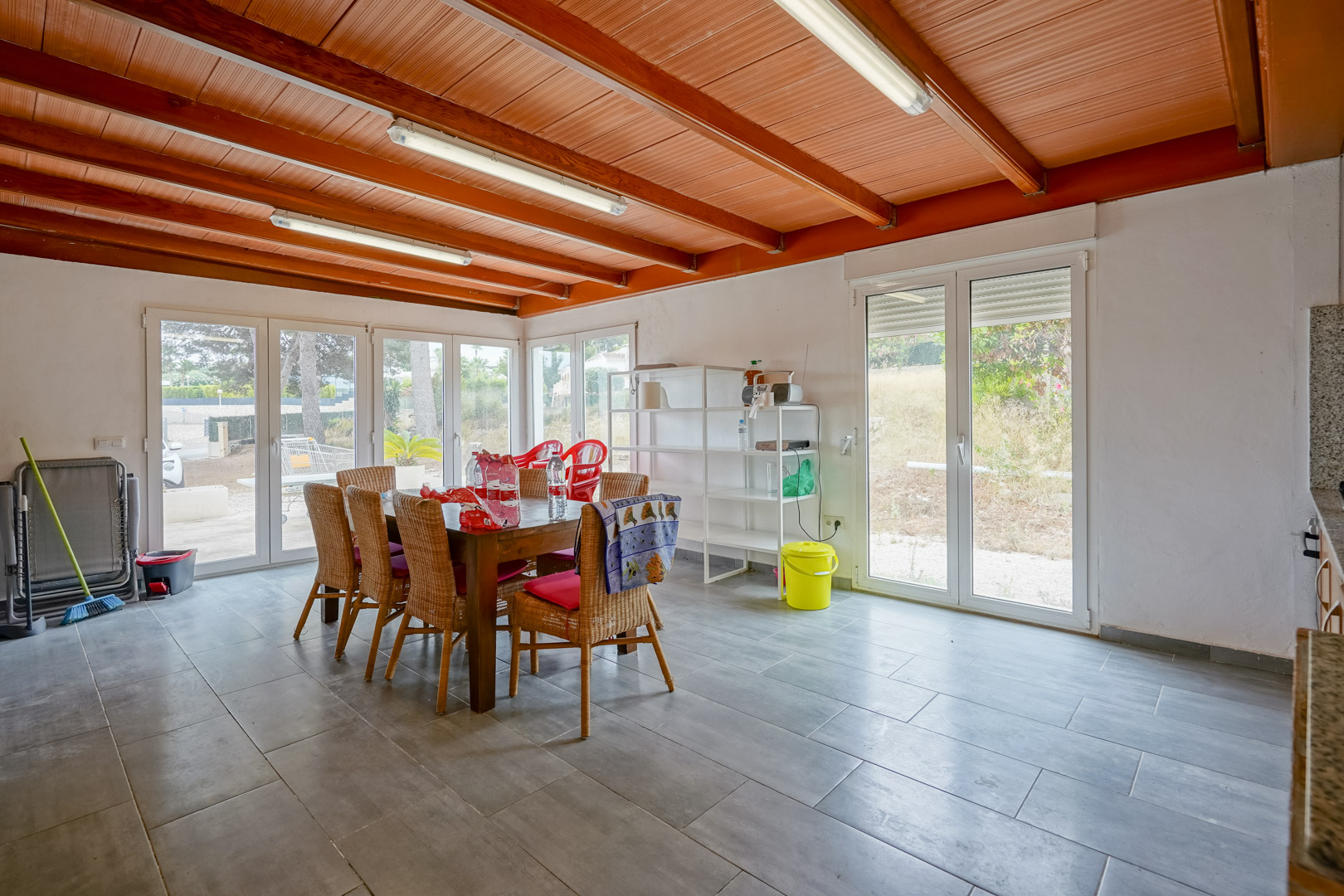 Comedor luminoso con techo de vigas de madera vistas, suelos de gres gris y amplios ventanales de PVC blanco con acceso directo al jardín exterior.