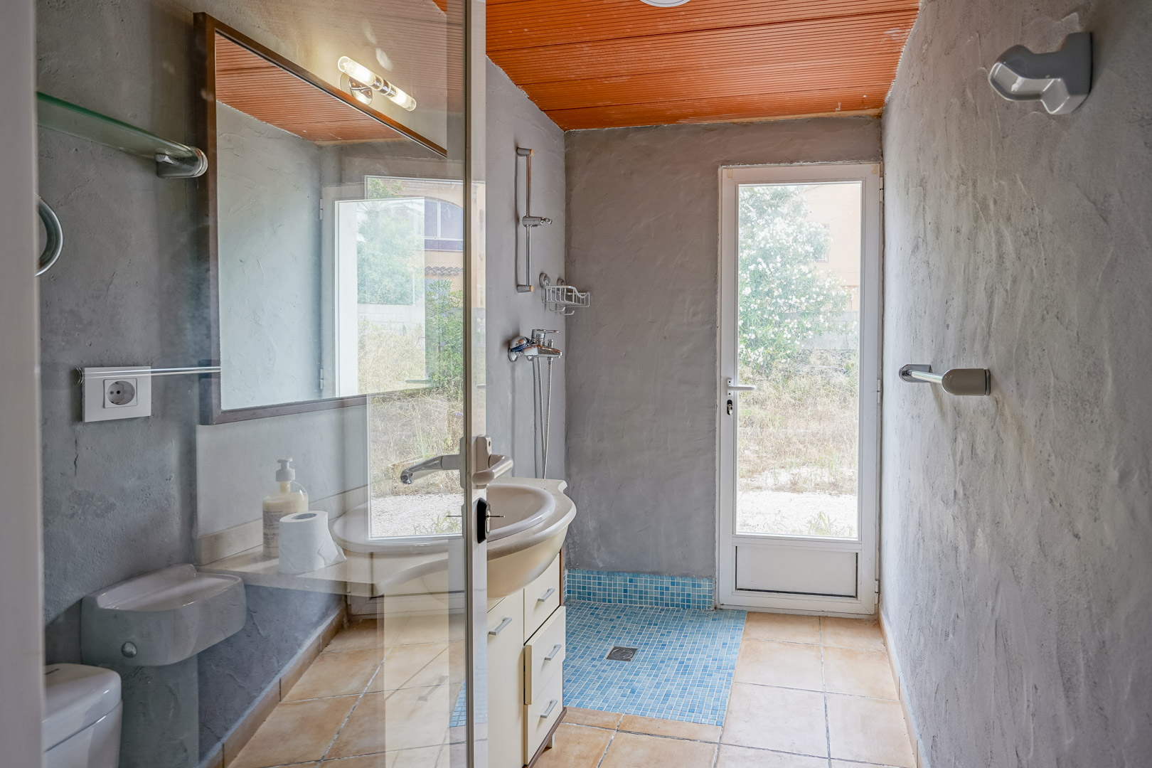 Cuarto de baño con paredes de cemento visto, techo de lamas naranjas y zona de ducha con gresite azul. Incluye acceso directo al exterior mediante una puerta acristalada.