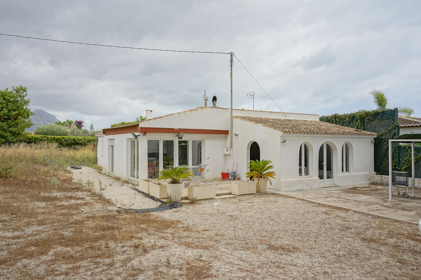 Fachada de la villa de estilo mediterráneo con arcos tradicionales, carpintería blanca y tejado de teja, situada sobre una extensa parcela llana con vistas al monte.