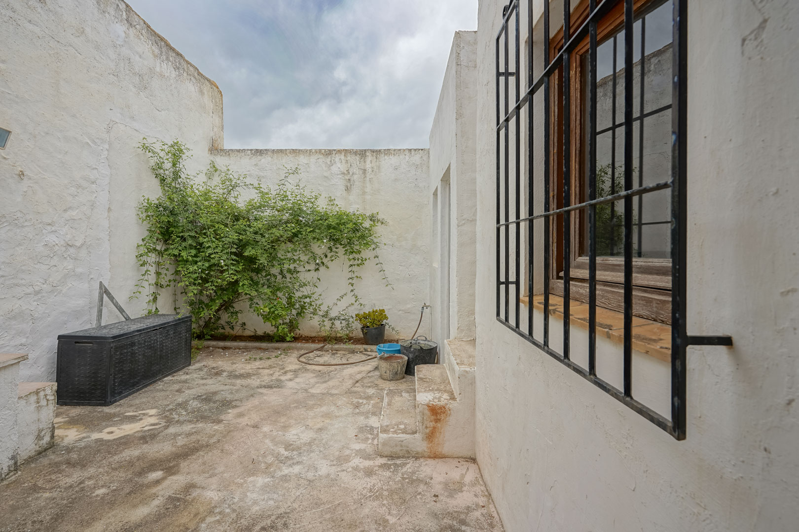 Patio interior con paredes encaladas, suelo de cemento, ventana con reja de hierro y zona de almacenaje exterior.