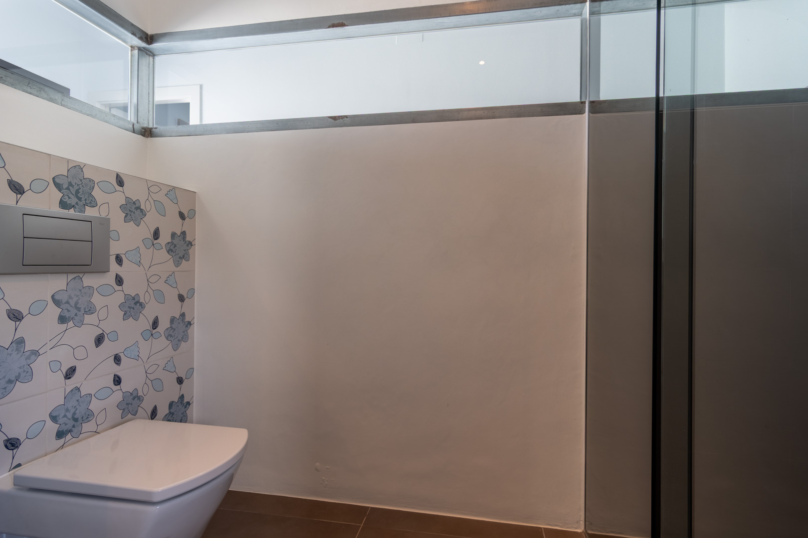 Baño moderno con inodoro suspendido, azulejos decorativos con motivos florales en tonos azules y un panel de vidrio superior que permite la entrada de luz natural.