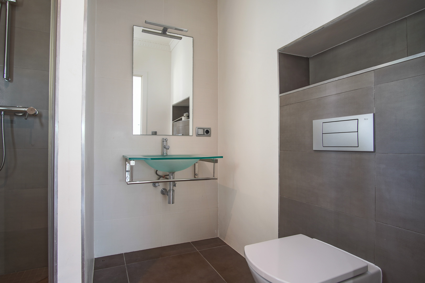 Baño moderno con inodoro suspendido, lavabo de cristal translúcido sobre soporte cromado y paredes revestidas en tonos neutros con hornacina integrada.