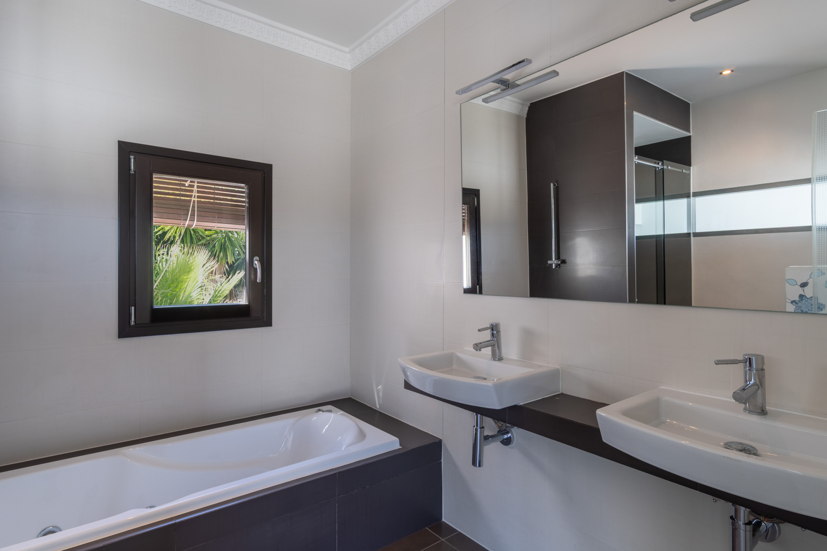 Baño moderno con lavabo doble sobre encimera oscura, bañera integrada y ventana con marco efecto madera que aporta luz natural y vistas al jardín.