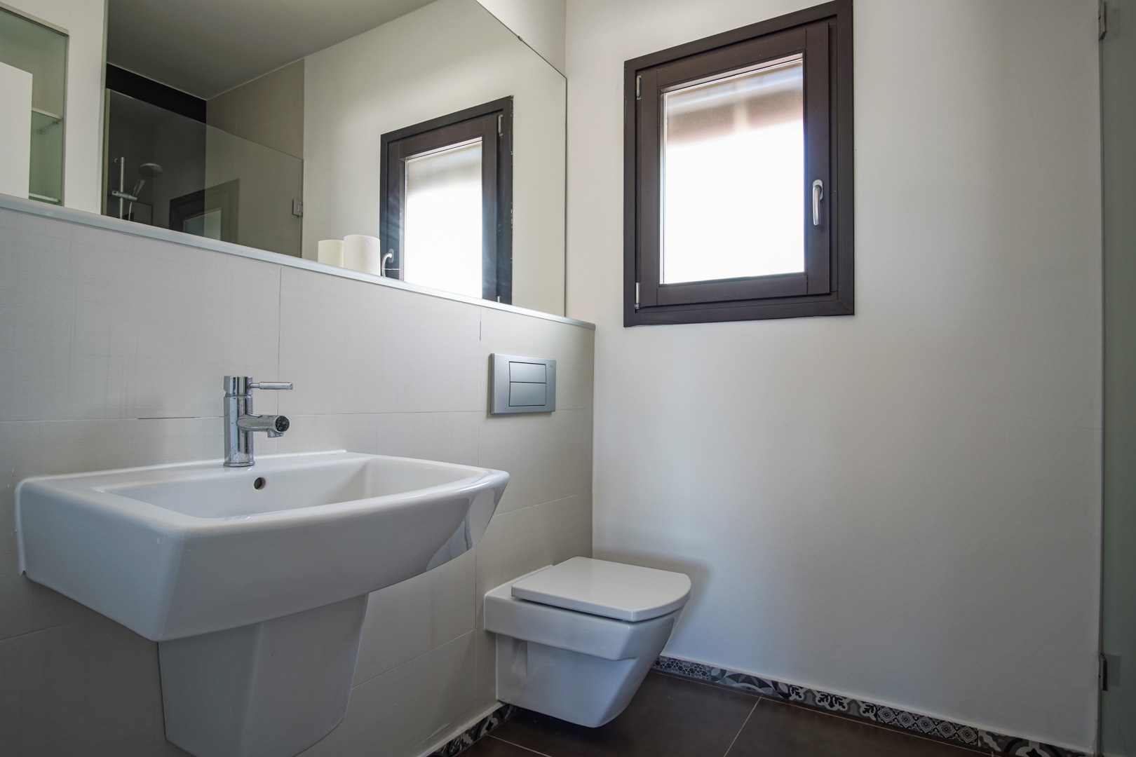 Baño moderno con sanitario suspendido, lavabo de cerámica blanca y ventana de aluminio. Incluye un gran espejo y suelos oscuros con cenefa decorativa de estilo hidráulico.