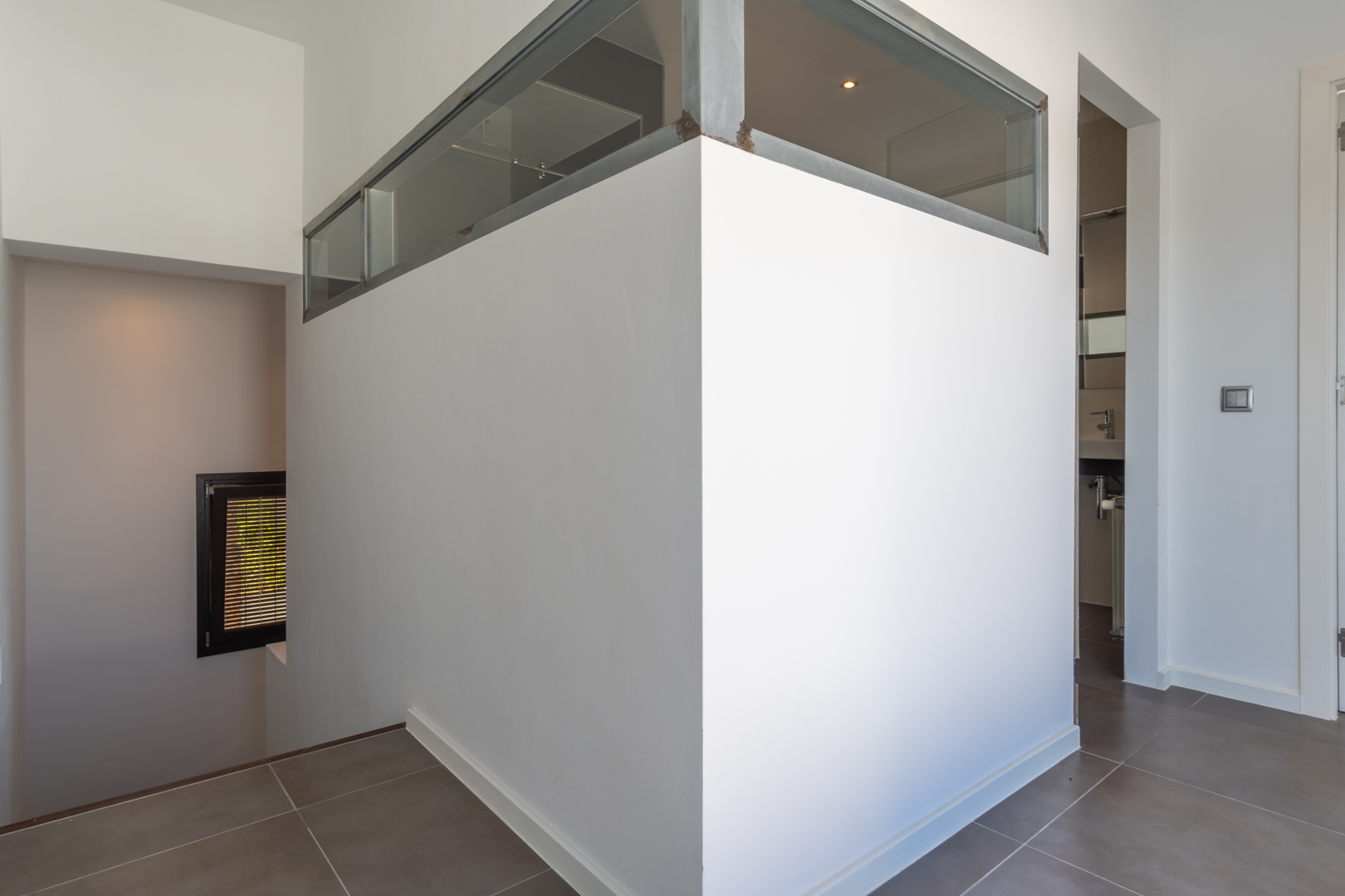 Distribuidor interior con suelos de gres, pared divisoria con cerramiento superior de cristal y metal, acceso a la escalera y vista parcial de un baño.