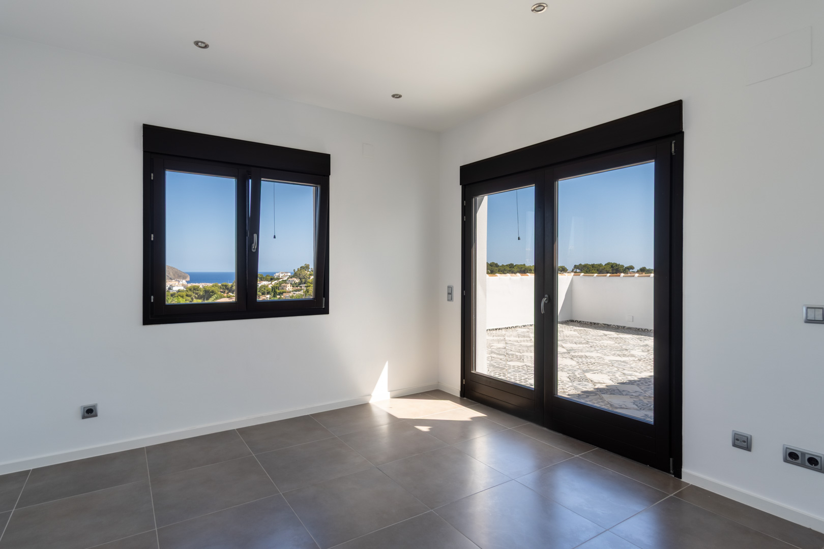Dormitorio con suelos cerámicos grises, carpintería exterior oscura y acceso directo a una terraza soleada con vistas despejadas al mar y la costa.