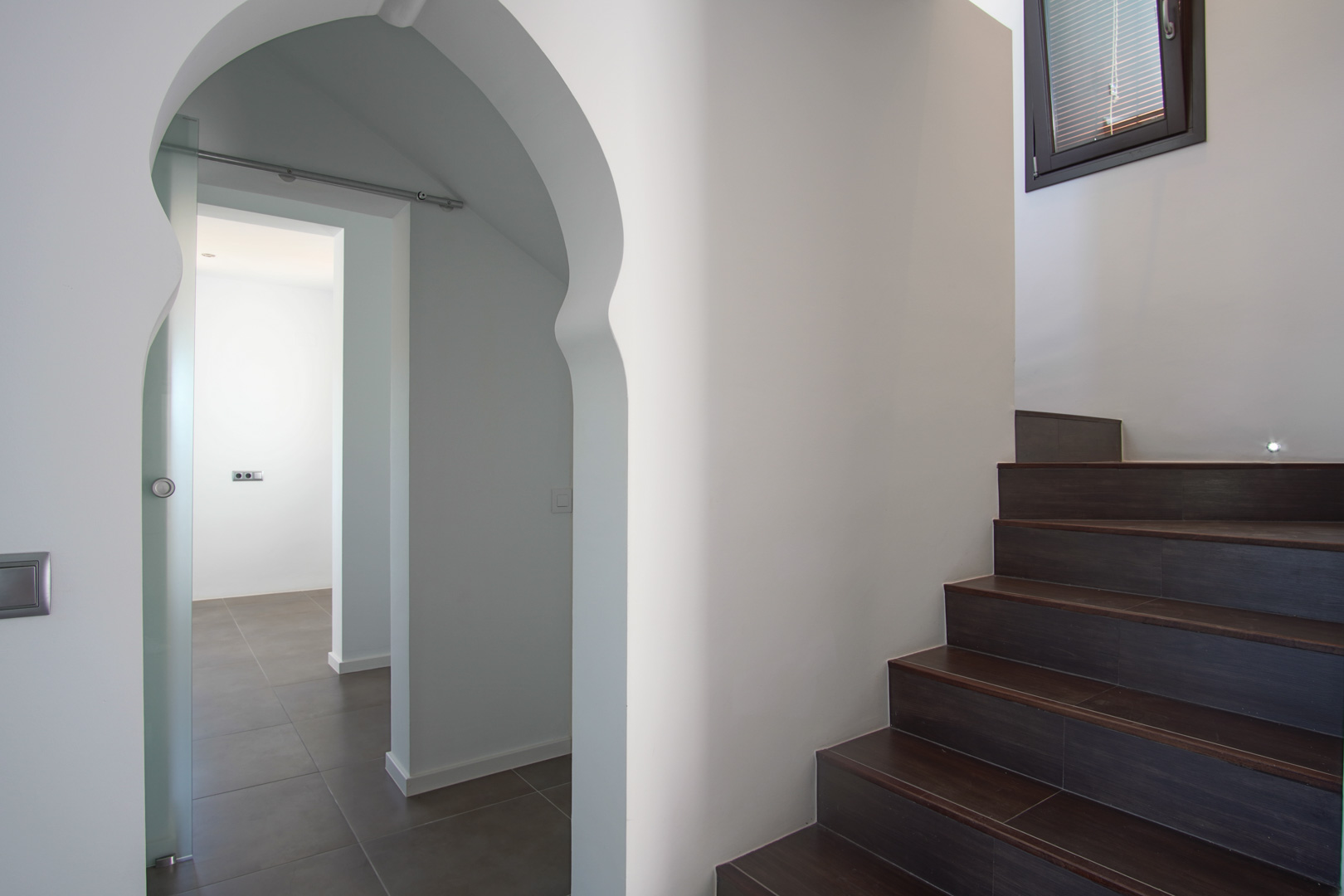Escalera interior con peldaños de gres oscuro e iluminación integrada, junto a un arco de estilo morisco que da paso a una estancia con puerta corredera de cristal.