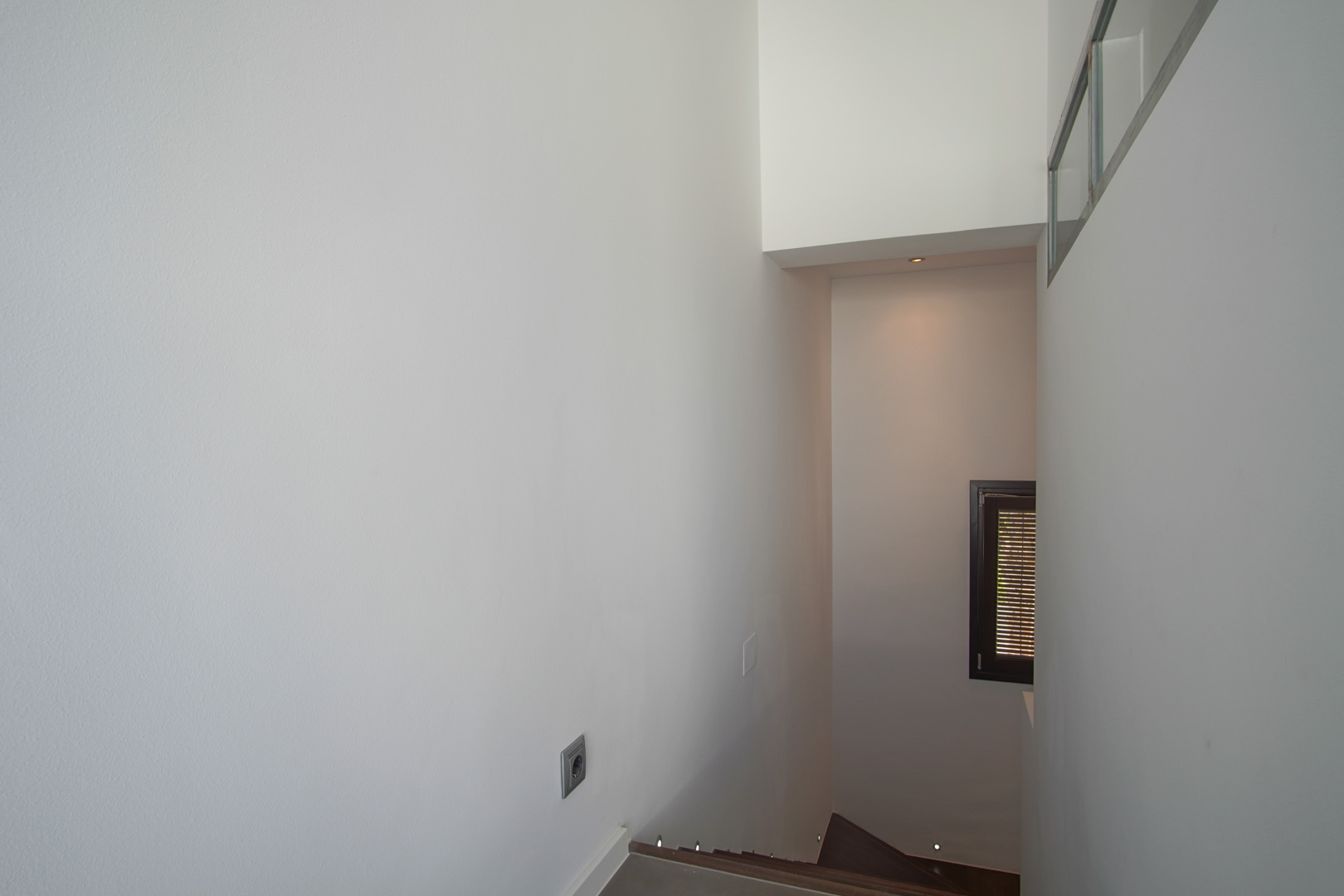 Escalera interior con peldaños oscuros e iluminación LED integrada en el suelo. Paredes blancas lisas y ventana con carpintería oscura y persiana de lamas.