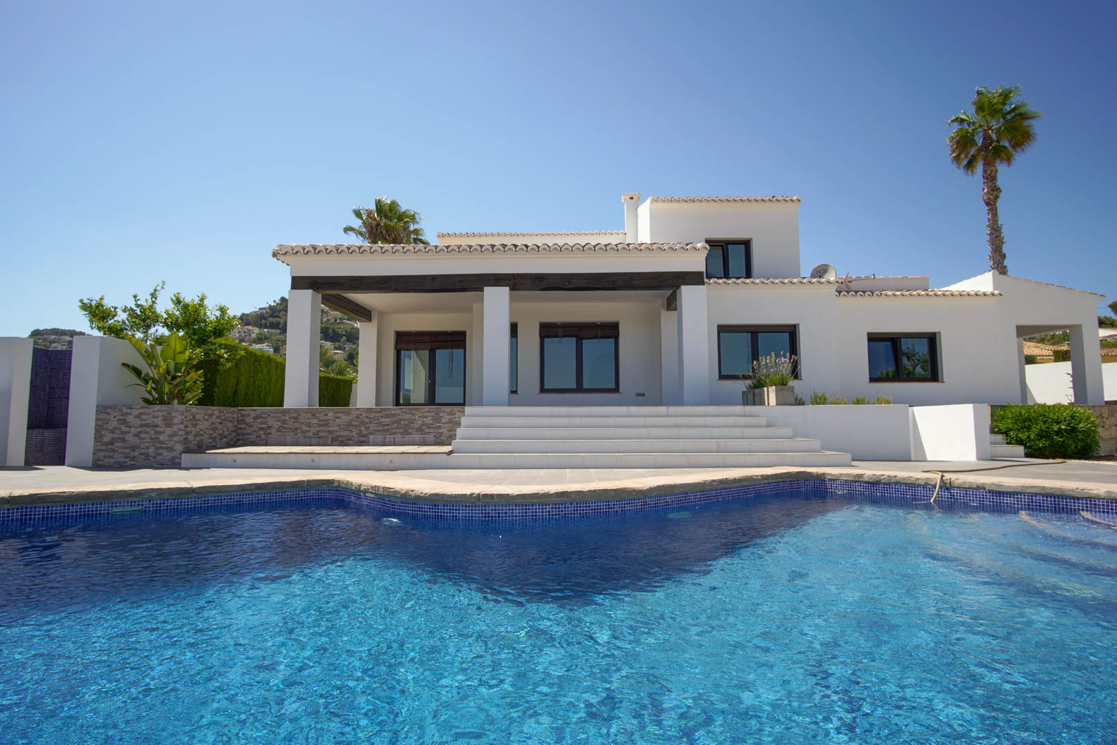 Fachada mediterránea blanca con piscina de mosaico azul. Destaca el porche cubierto con vigas de madera oscura y las amplias escaleras que acceden a la vivienda.