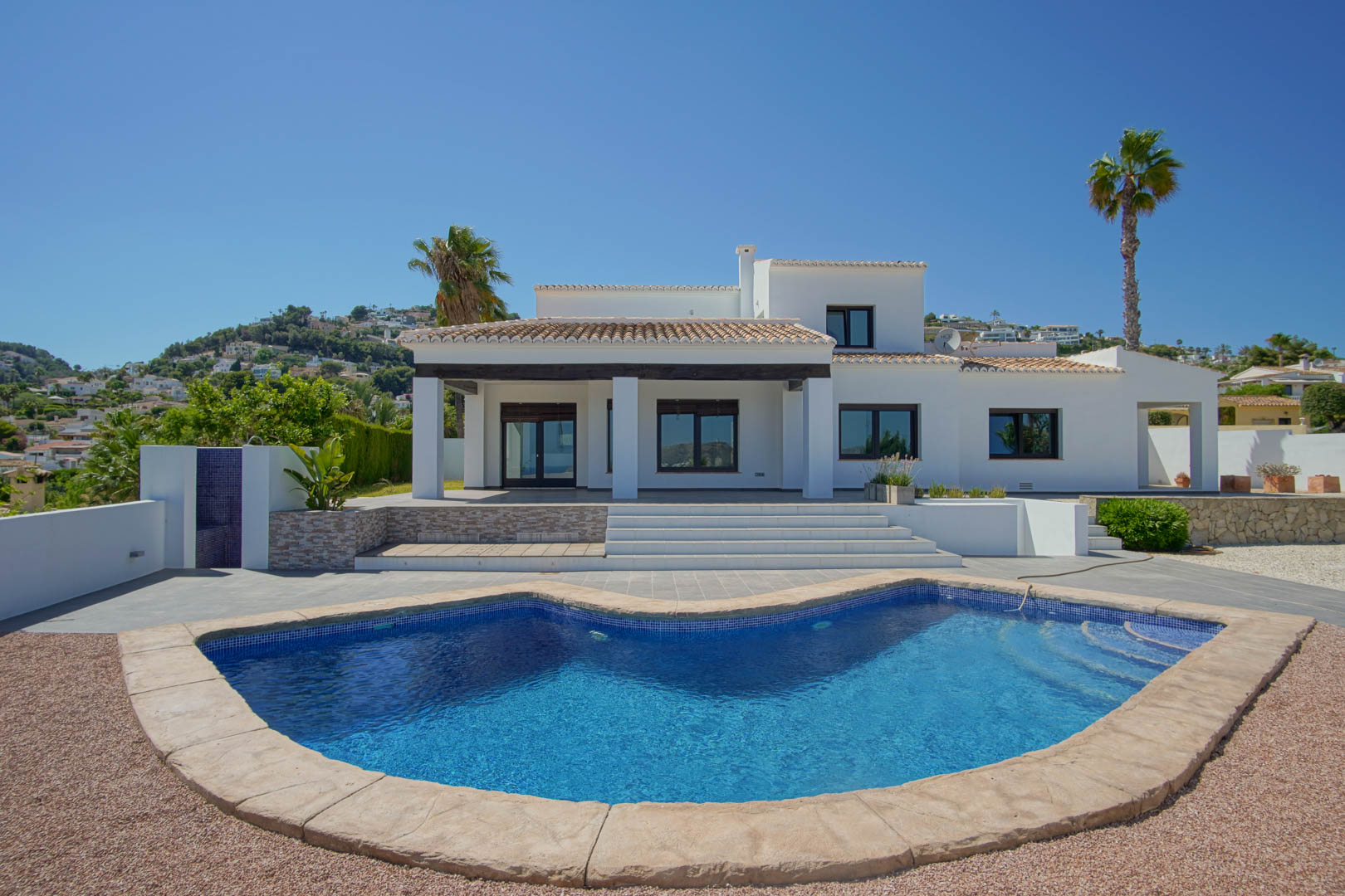Fachada principal de la villa con piscina de diseño curvo, porche cubierto con vigas de madera oscura y amplias terrazas blancas integradas en el entorno.