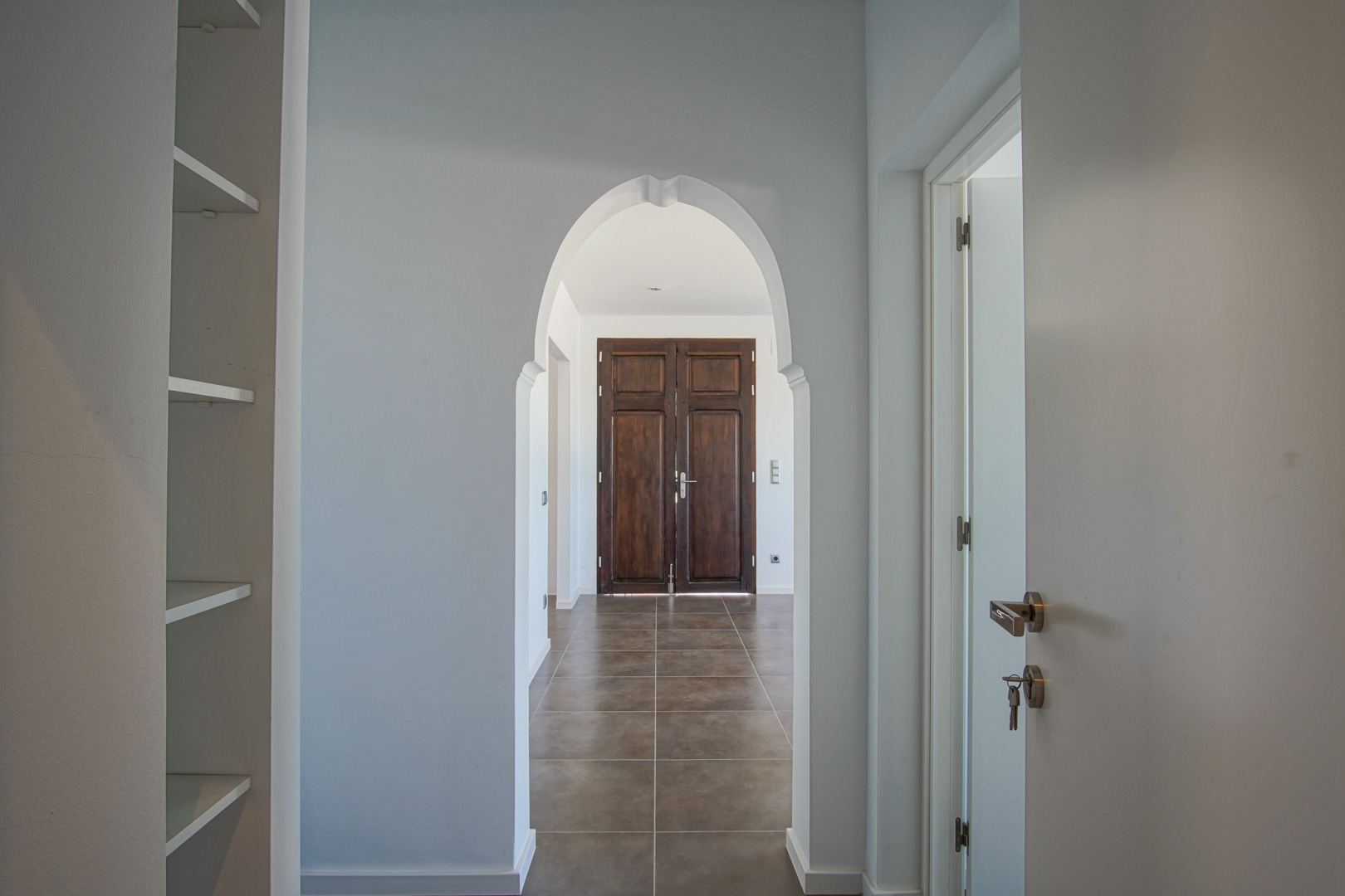 Pasillo interior con arco de estilo mediterráneo que enmarca una puerta doble de madera oscura restaurada sobre suelos cerámicos de tonos tierra.