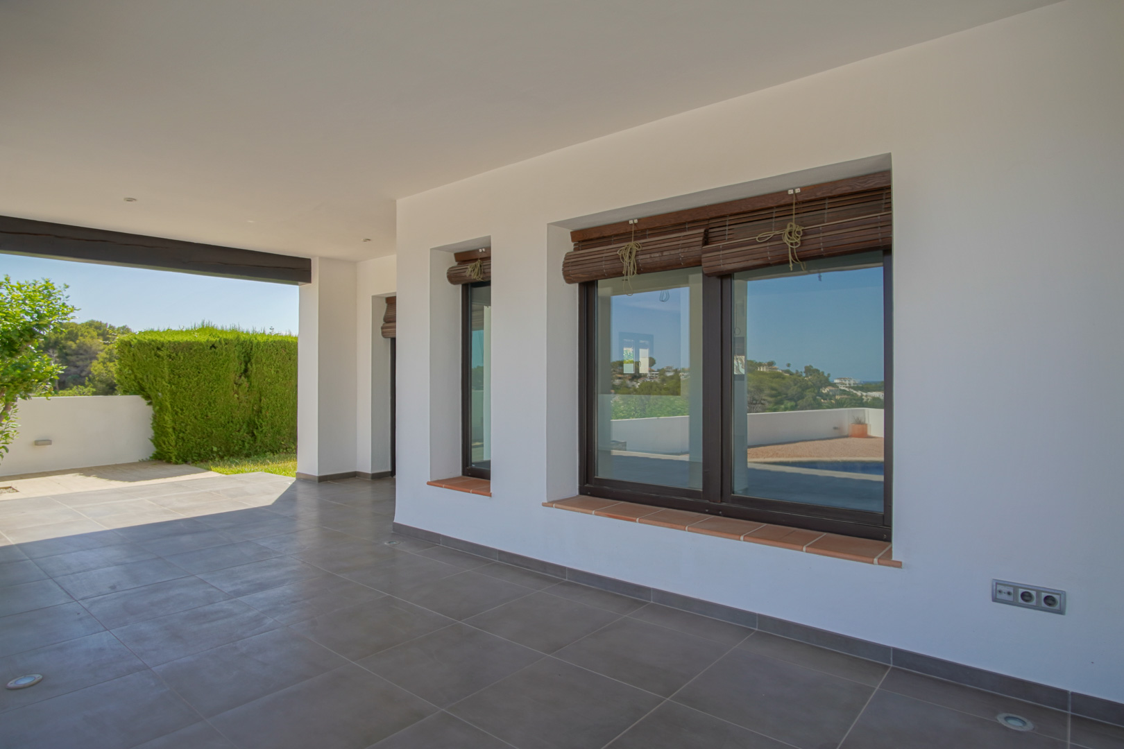 Porche cubierto con suelo de gres gris, paredes blancas y ventanales de aluminio efecto madera con persianas de esterilla y alféizares de terracota.