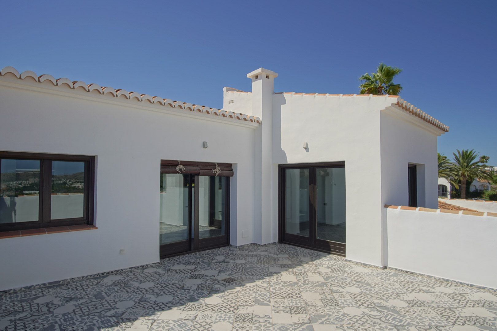 Terraza solárium con suelo de gres porcelánico decorado, carpintería exterior de aluminio efecto madera y arquitectura blanca mediterránea.