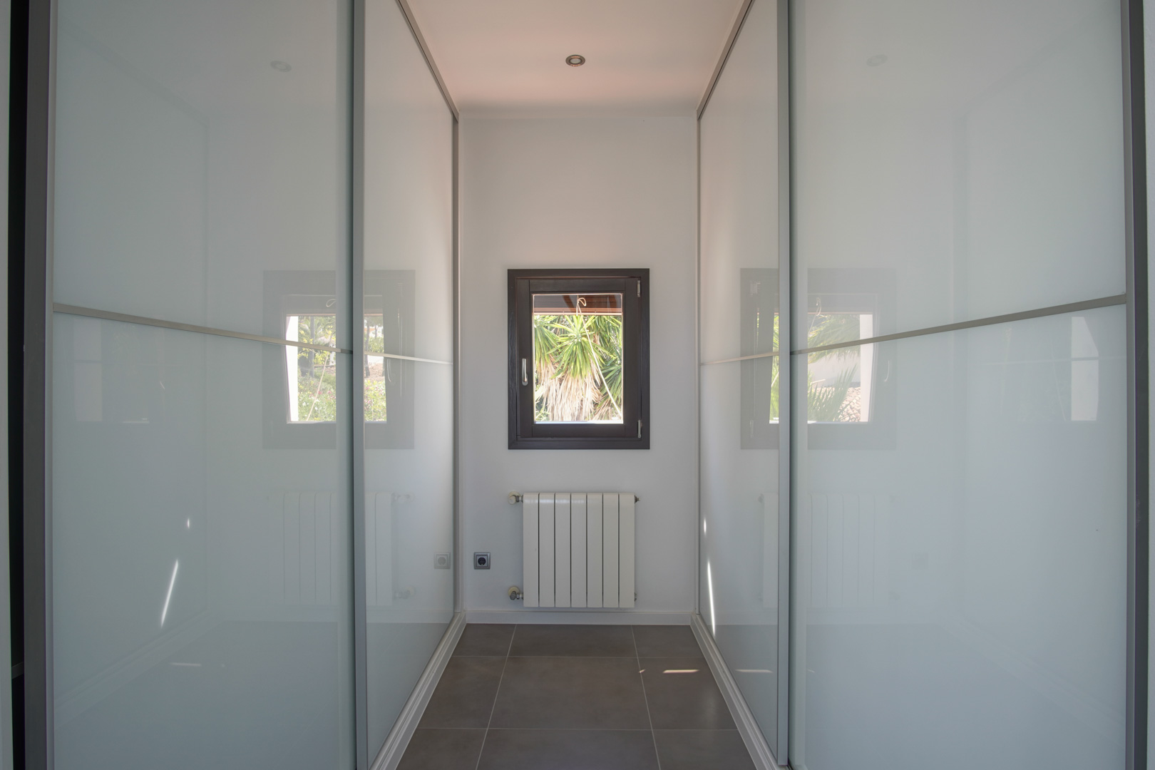 Vestidor con armarios empotrados de puertas correderas en cristal traslúcido, radiador bajo ventana de marco oscuro y suelos cerámicos.