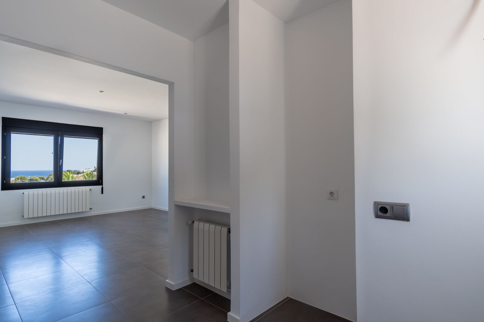 Vista desde el distribuidor hacia una estancia con suelos cerámicos, radiadores de pared y ventana de aluminio negro con vistas despejadas al mar.