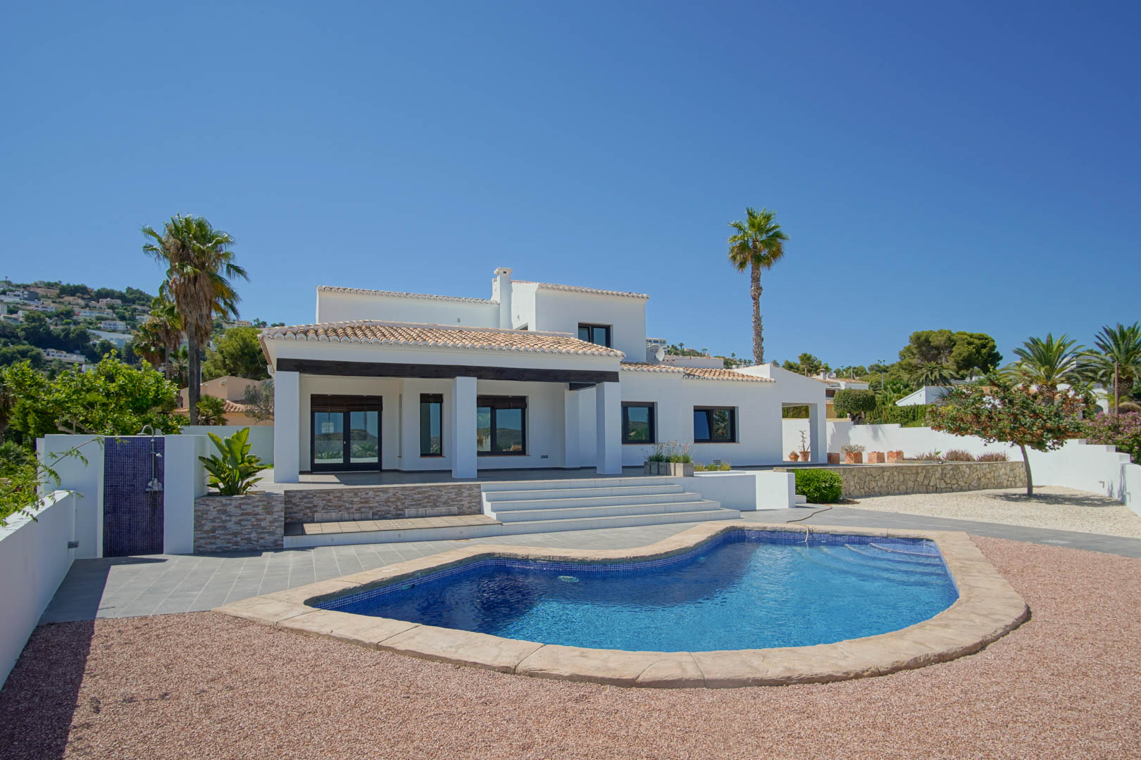 Vista exterior de la villa con piscina de diseño orgánico, rodeada de zonas de grava y una terraza cubierta con vigas de madera oscura y columnas blancas.