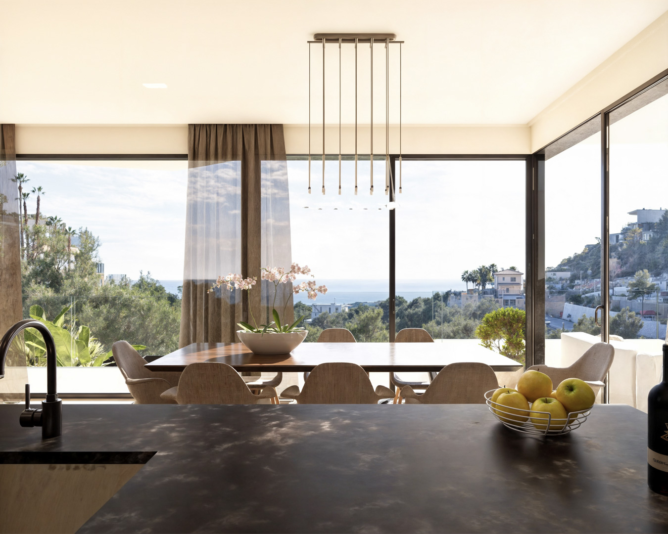 Comedor integrado con cocina, mesa de madera, lámpara de diseño y amplios ventanales con vistas panorámicas al mar Mediterráneo y la costa de Benissa.