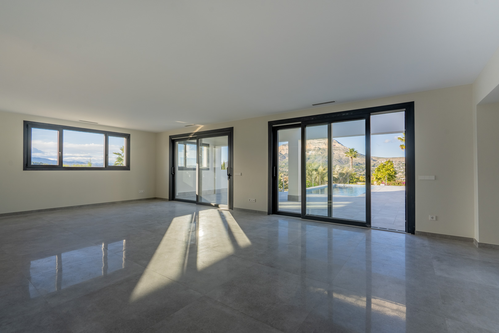 Amplio salón con suelo de baldosas grises y grandes ventanales que ofrecen vistas a la piscina y al paisaje.
