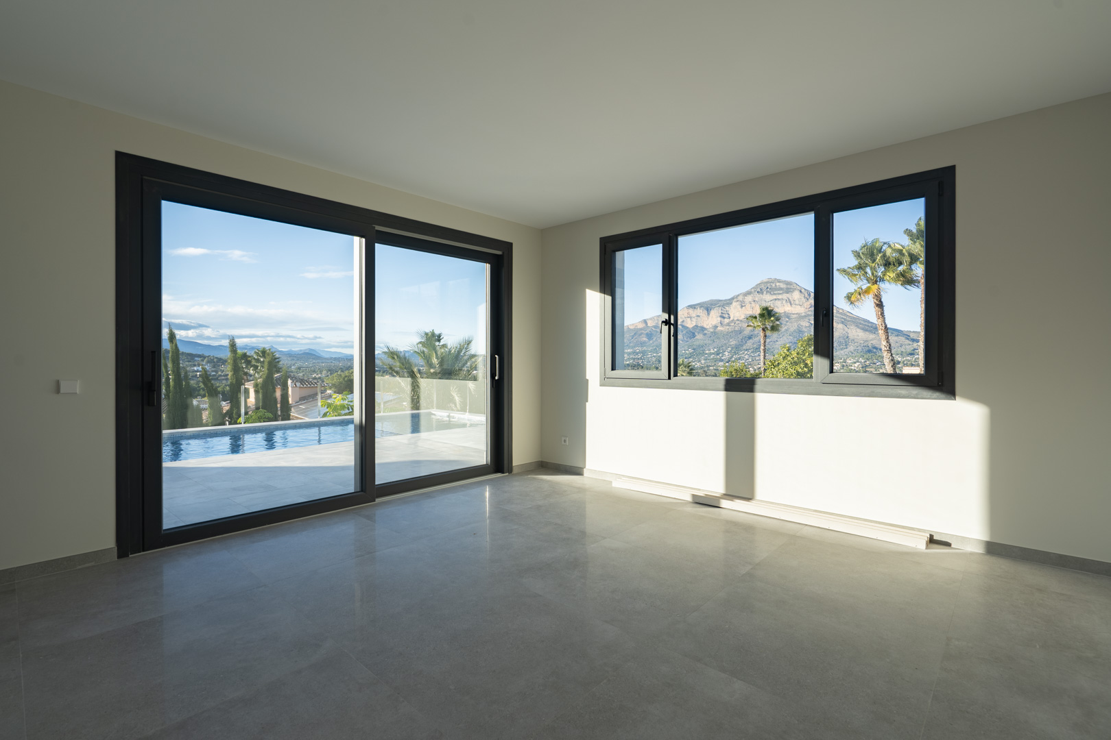 Espacioso salón con suelo de baldosas grises y grandes ventanales con vistas a la piscina y al paisaje montañoso.