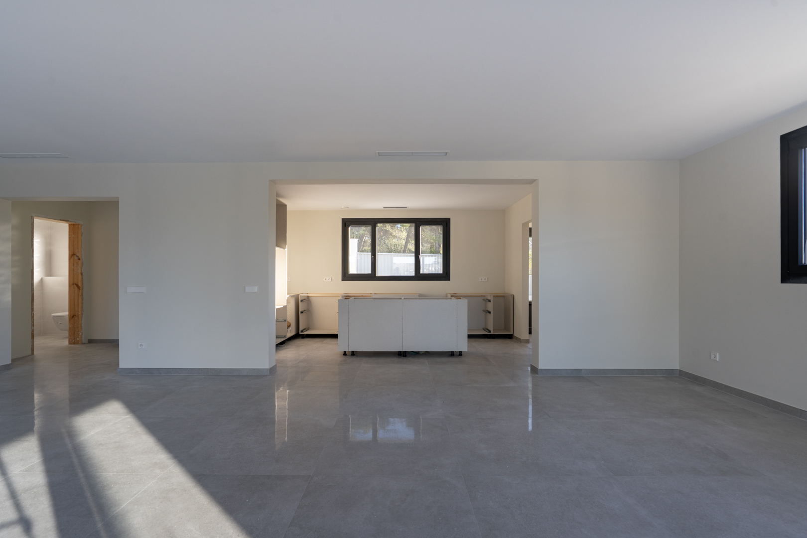 Interior diáfano con suelo de gres gris. Cocina en obra con muebles bajos blancos y ventana con carpintería negra.