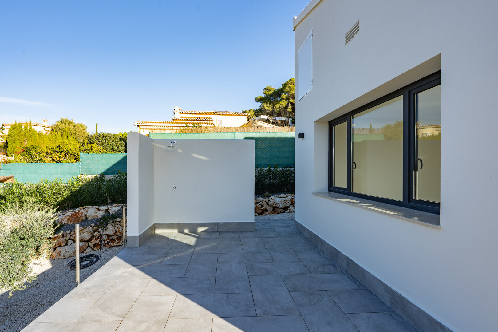 Patio con ducha exterior y baldosas grises, junto a la pared blanca con ventana de doble acristalamiento.