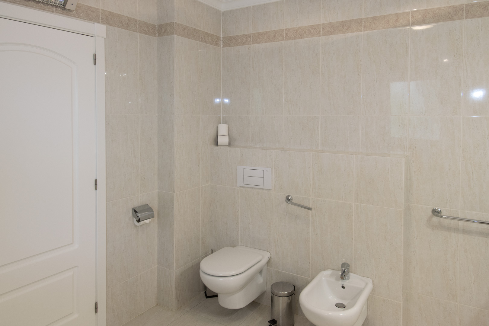 Cuarto de baño con azulejos en tonos crema, inodoro suspendido, bidé y barras de seguridad cromadas integradas en las paredes alicatadas.