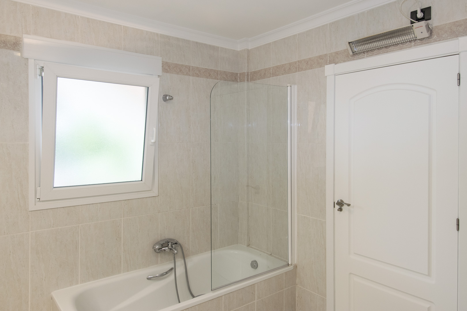 Cuarto de baño con bañera y mampara de cristal, paredes alicatadas con cenefa ornamental, ventana de PVC con vidrio translúcido y calefactor eléctrico de pared.