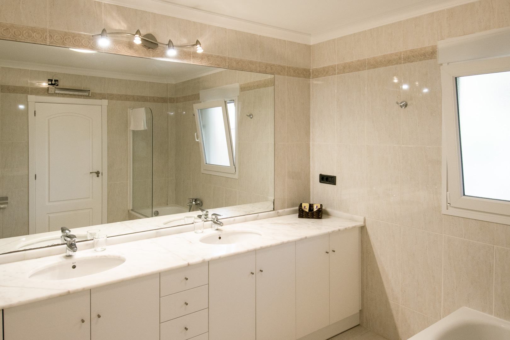 Cuarto de baño con encimera de mármol blanco, lavabo de doble seno y un gran espejo de pared. Incluye revestimiento cerámico en tonos crema y una ventana para luz natural.