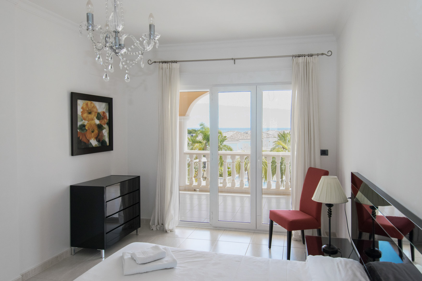 Dormitorio con acceso a terraza privada, mobiliario moderno y vistas al mar. Destaca la lámpara de araña de cristal y el suelo de gres porcelánico de tonos claros.