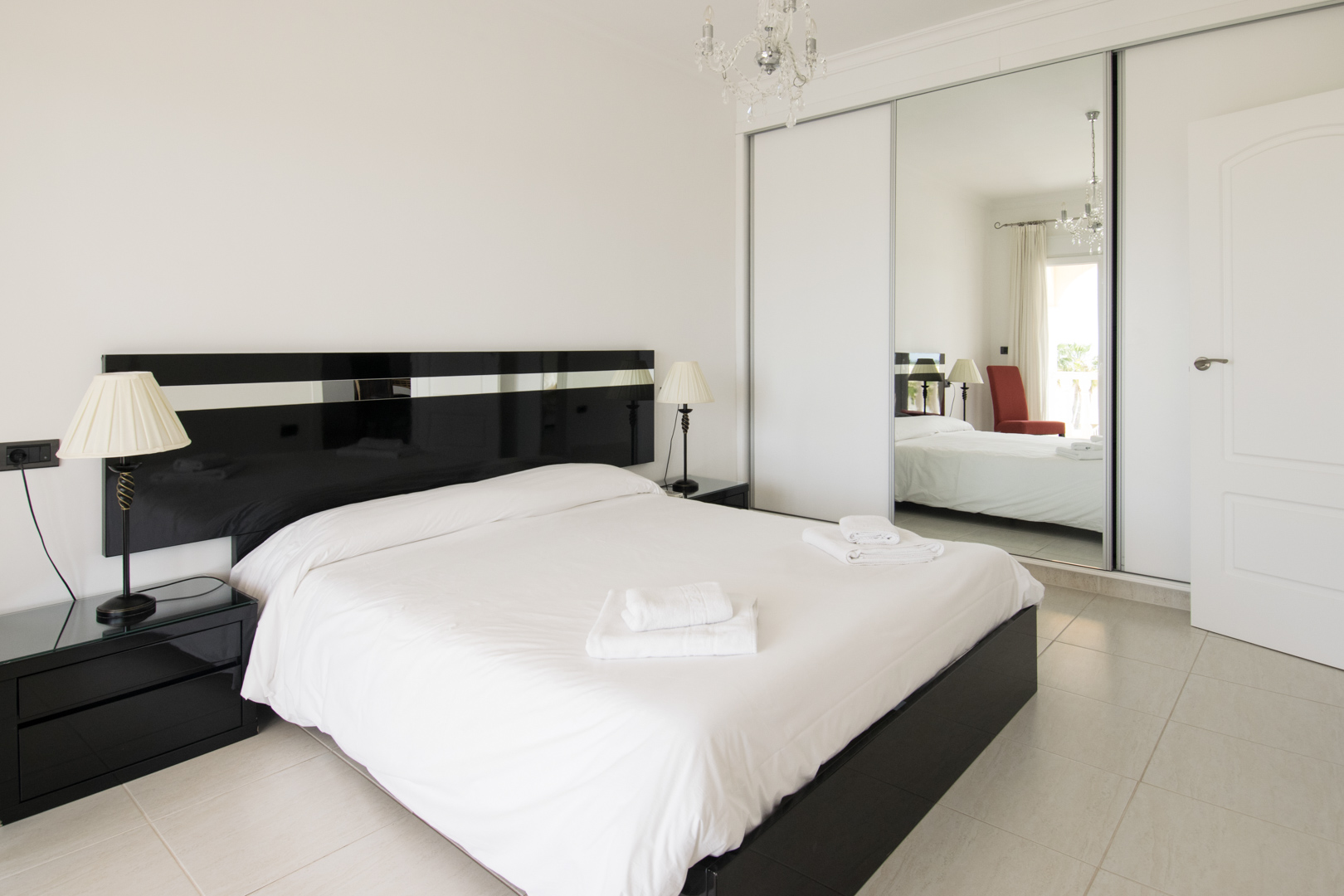 Dormitorio principal con cama doble, cabecero negro brillante, mesitas de noche a juego y un amplio armario empotrado con puertas correderas de espejo y lámpara de araña.