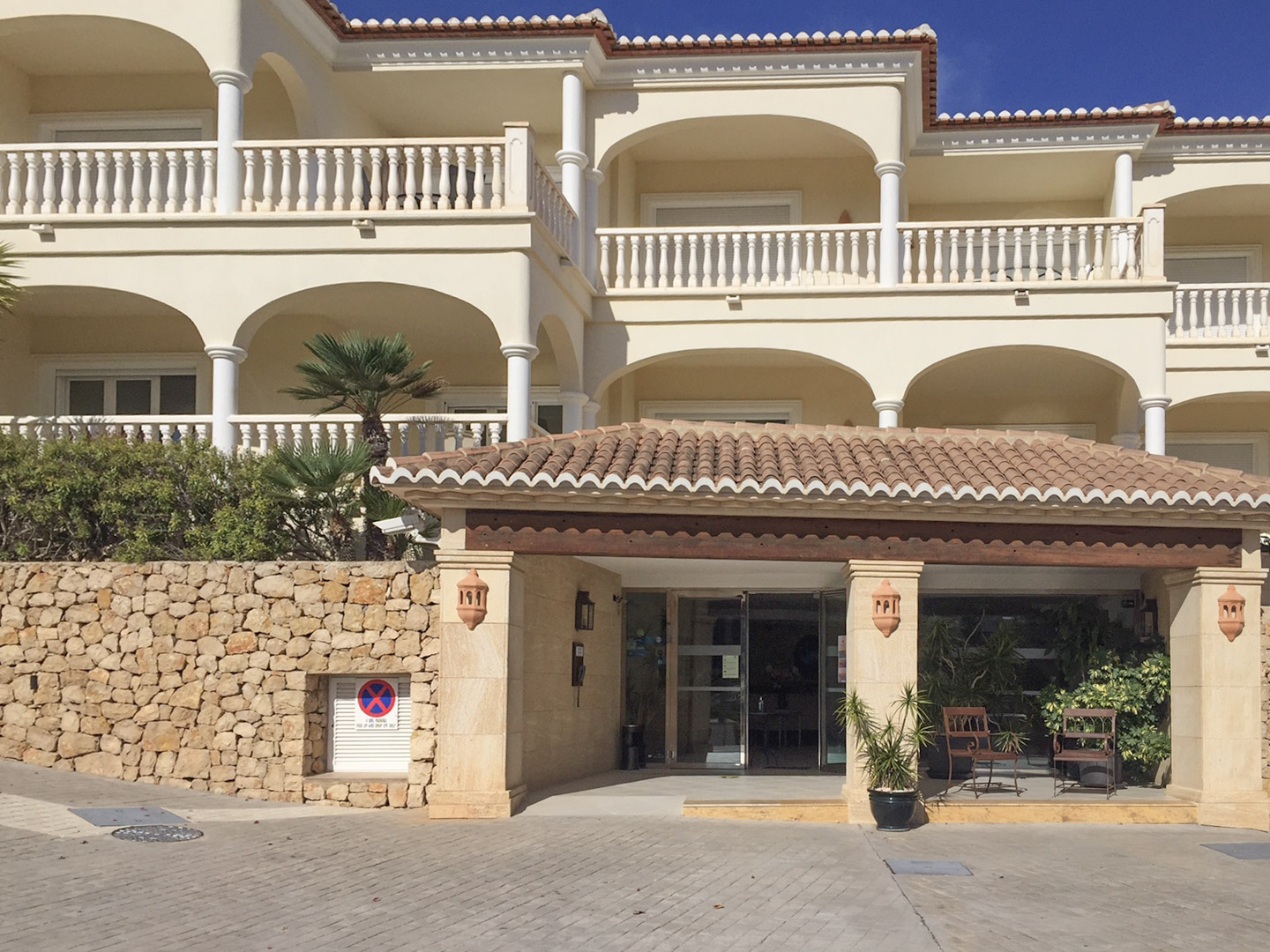 Entrada principal del complejo con porche de vigas de madera, muros de piedra natural y fachadas mediterráneas con arcos y balaustradas blancas.