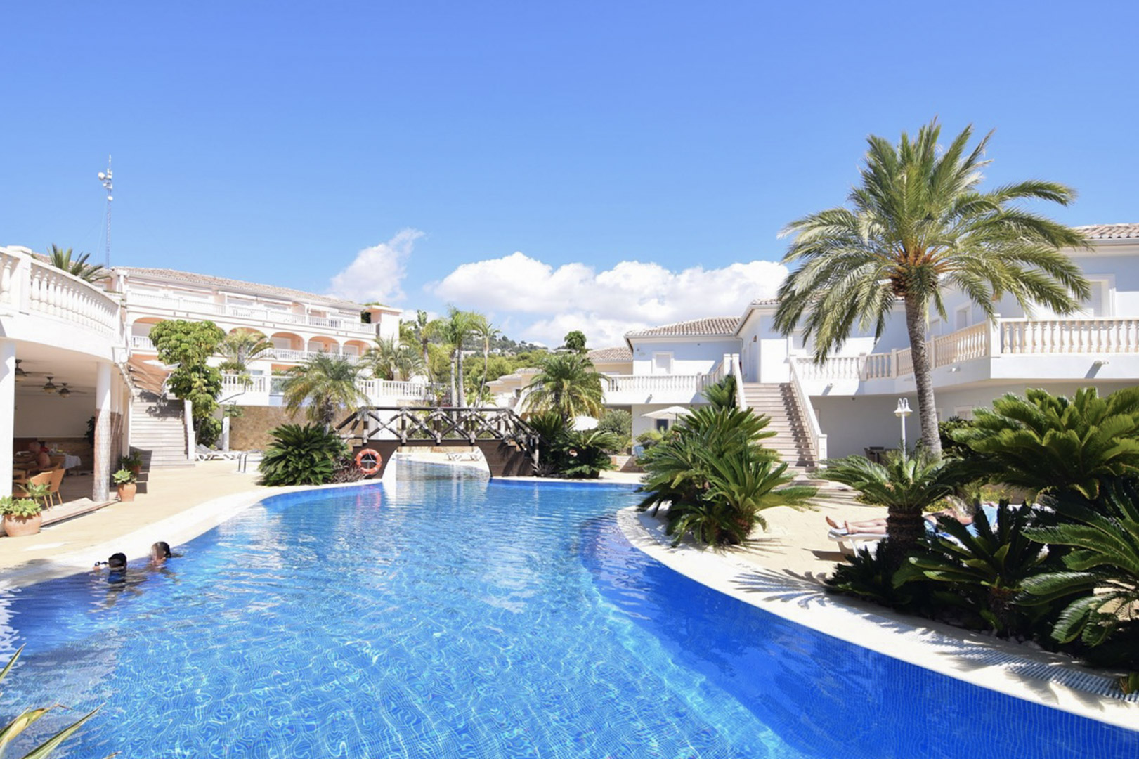 Piscina comunitaria tipo laguna con puente de madera, rodeada de vegetación mediterránea y edificios blancos de estilo clásico en Benissa Costa.