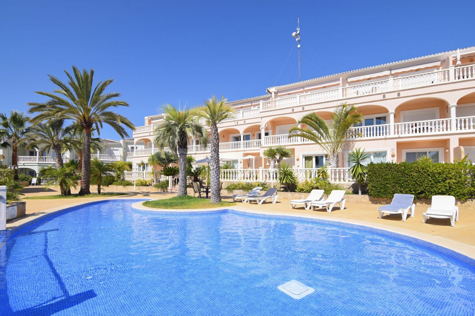 Piscina comunitaria tipo laguna rodeada de palmeras y zonas de solárium con tumbonas, frente a la fachada de estilo mediterráneo con amplias terrazas y balaustradas blancas.