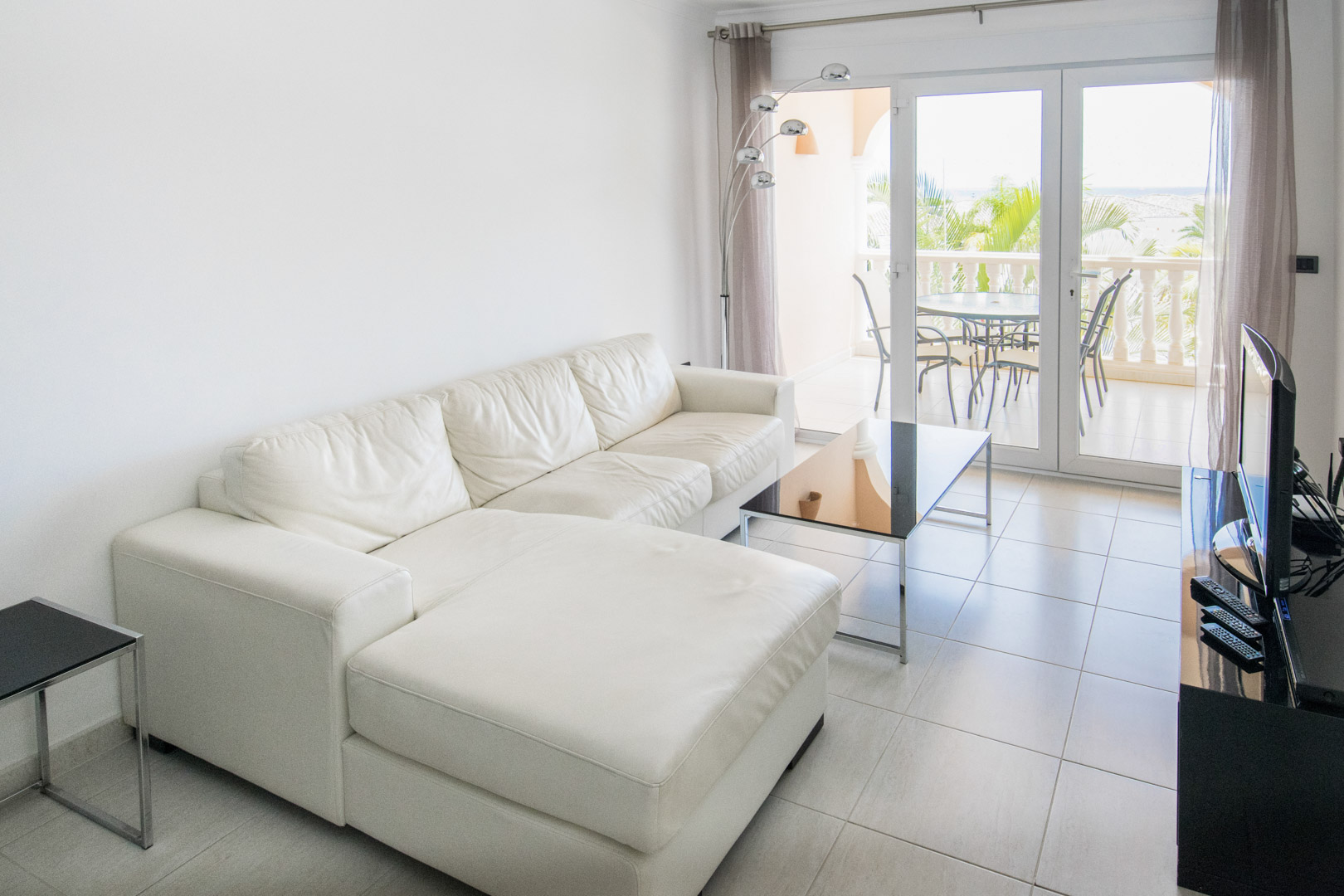 Salón luminoso con sofá rinconera de piel blanca, suelos de gres claro y acceso directo a una terraza amueblada con vistas a las palmeras y el mar.