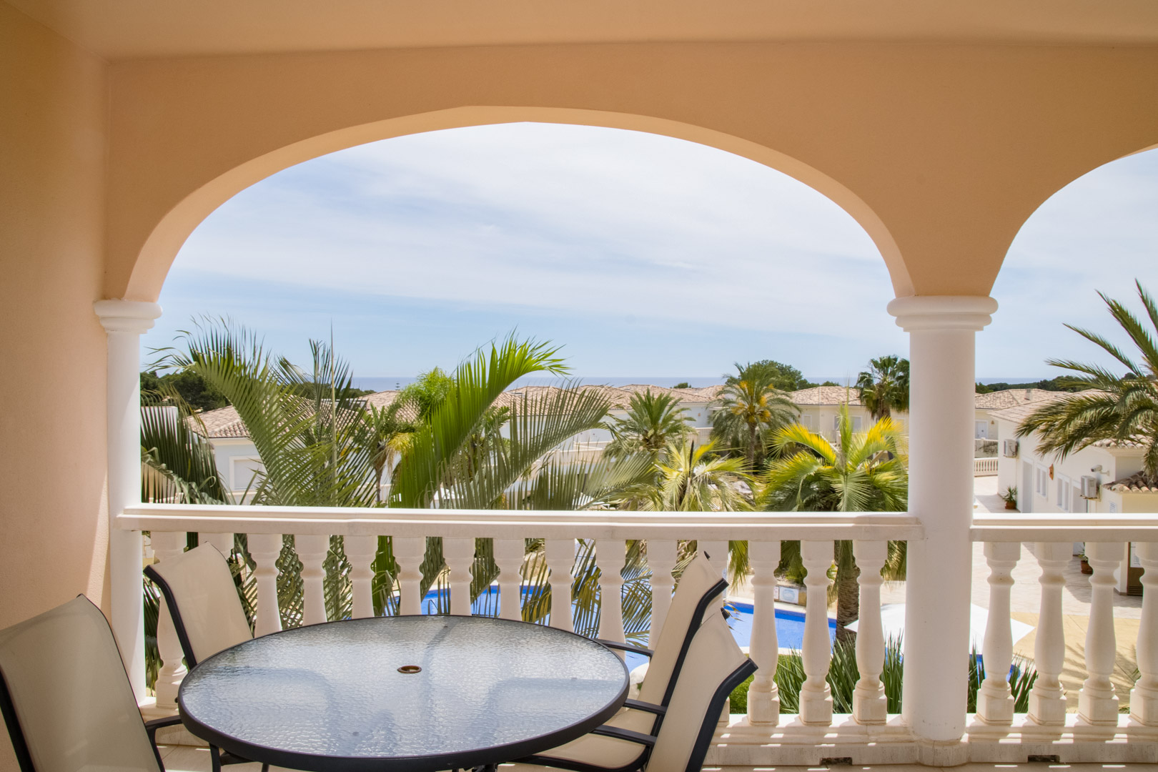 Terraza cubierta con arcos y balaustrada clásica, equipada con mesa de cristal. Ofrece vistas a los jardines mediterráneos, la piscina tipo laguna y el mar.